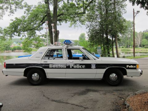 1991 Ford LTD Crown Victoria - restored Boston Police white-and-black Ford sedan shown in side profile with a roof lightbar, parked near a tree-lined pond.