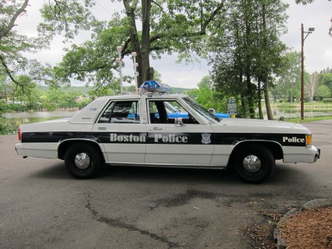 1991 Ford LTD Crown Victoria - Driver-side view of a white-and-black Boston Police Ford sedan with roof lights, parked on a paved lot by trees and water.