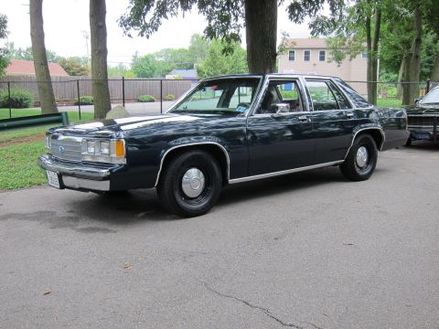 1991 Ford LTD Crown Victoria - dark navy-blue four-door Ford sedan shown front three-quarter with steel wheels, parked on a paved lot behind a chain-link fence.