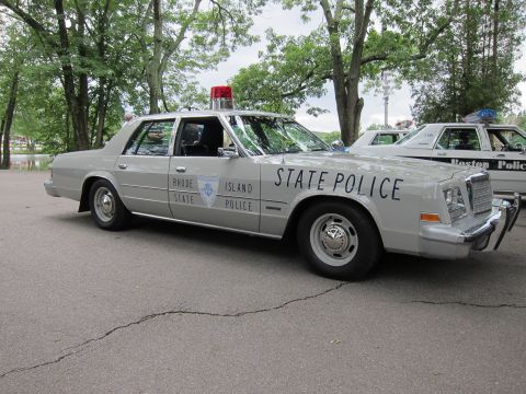 1991 Ford LTD Crown Victoria - silver Rhode Island State Police sedan with a single red roof beacon, parked beside a Boston Police car on a paved lot.