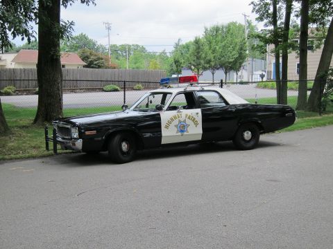 1991 Ford LTD Crown Victoria - Front three-quarter view of a black-and-white Highway Patrol sedan with a red roof beacon and push bar, parked by a fence.