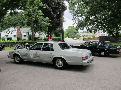 1991 Ford LTD Crown Victoria - silver Rhode Island State Police sedan parked next to a dark four-door sedan with motorcycles behind, in a residential lot.