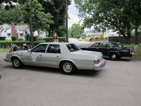 1991 Ford LTD Crown Victoria - silver Rhode Island State Police sedan parked beside a dark four-door sedan with motorcycles and houses behind.