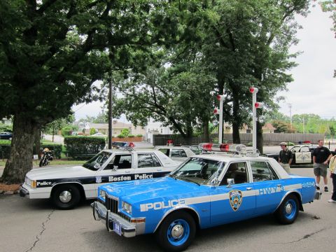1991 Ford LTD Crown Victoria - blue NYPD police sedan parked beside a white-and-black Boston Police car under shade trees, with people standing nearby.