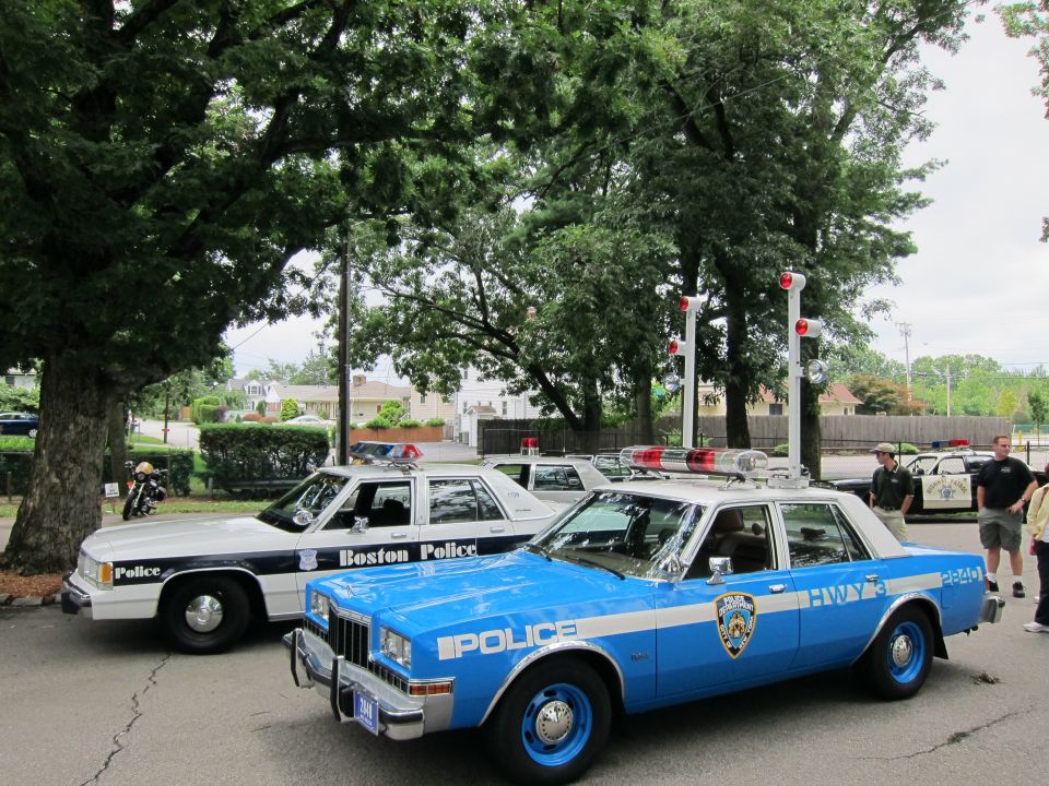 Blue NYPD police sedan and white Boston Police car parked under trees