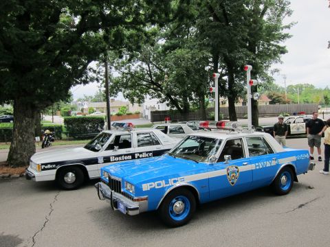 1991 Ford LTD Crown Victoria - Front angle of a blue NYPD police sedan parked beside a white-and-black Boston Police car under trees on a paved lot.