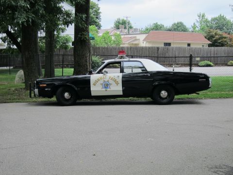 1991 Ford LTD Crown Victoria - Driver-side profile of a black-and-white Highway Patrol sedan with a white roof, red beacon and push bar, parked by a fence.