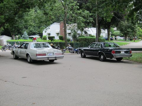 1991 Ford LTD Crown Victoria - Rear view of a silver State Police sedan and a dark four-door sedan parked across a paved lot with houses and motorcycles behind.