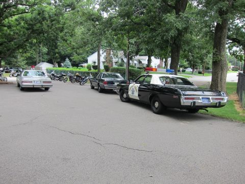 1991 Ford LTD Crown Victoria - row of vintage police cars parked along a tree-lined lot, led at the rear by a black-and-white Highway Patrol sedan.