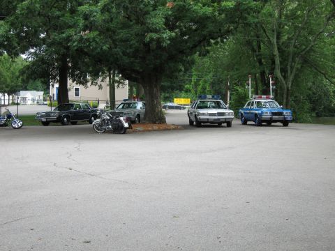 1991 Ford LTD Crown Victoria - group of vintage police cars and a parked motorcycle gathered around a large tree on an open paved lot.