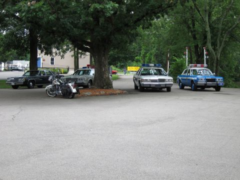 1991 Ford LTD Crown Victoria - Several vintage police cars and a motorcycle parked around a large tree on an open paved lot near houses.