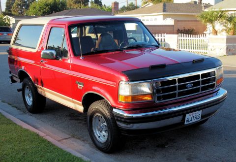 1994 Ford Bronco - This red Ford Bronco with a tan top is parked on a street, showing its front grille and hood cover.