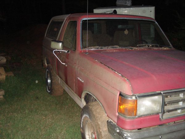 Red Ford Bronco parked in a grassy yard at night. The vehicle appears dusty and weathered