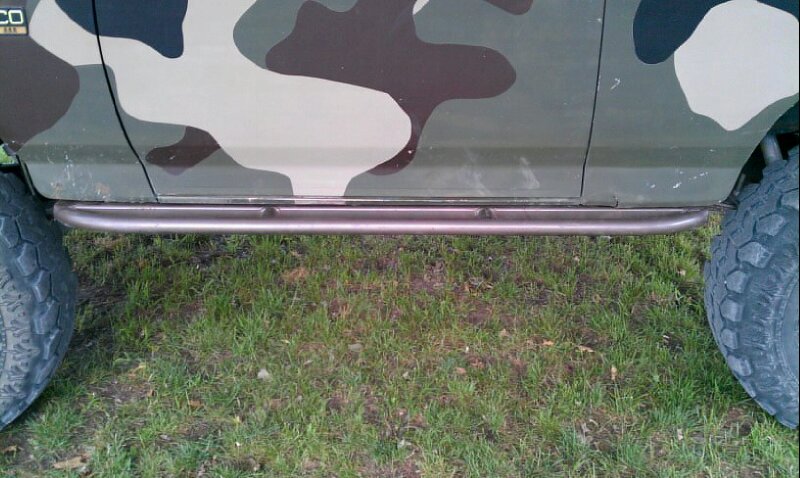 Side view of a 1995 Ford Bronco with camouflage paint and tubular rock sliders. Large off-road tires are visible