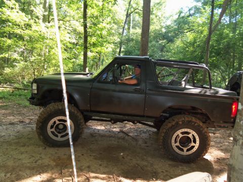 1987 Ford F-250 - Driver in a modified Ford Bronco navigates a wooded trail with large off-road tires.