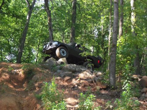 1987 Ford F-250 - A black truck with modified suspension climbs over rocks on a wooded trail.