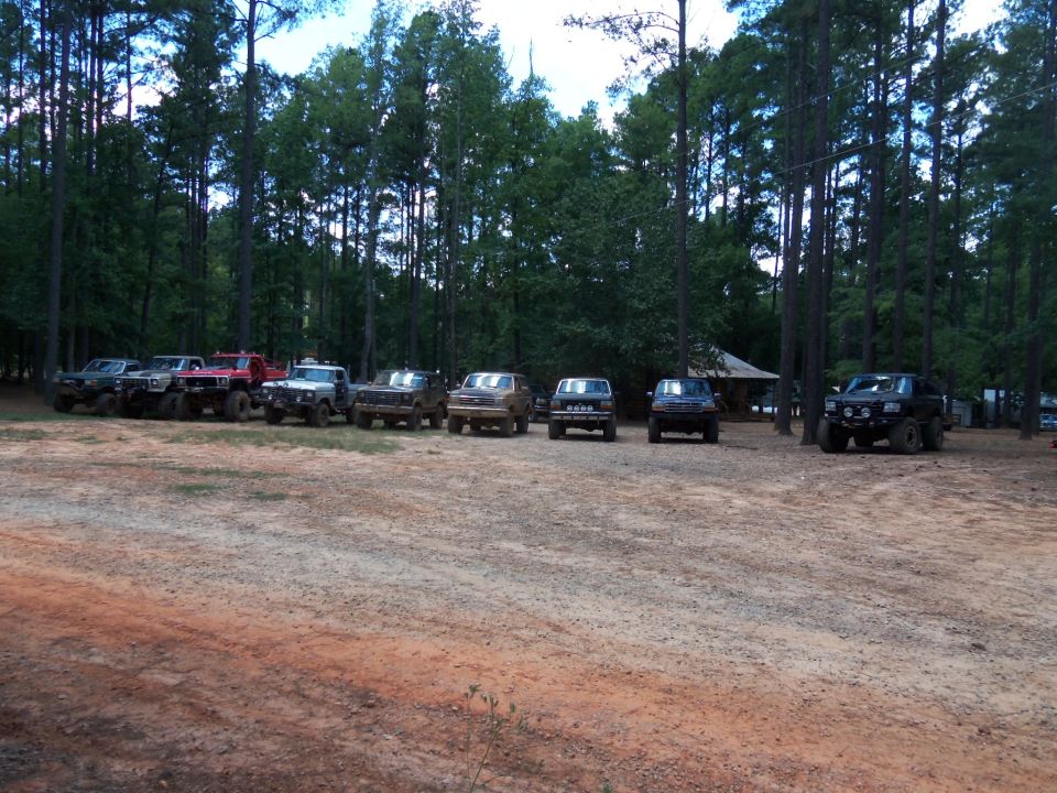 Several off-road trucks parked in a dirt lot with trees in the background