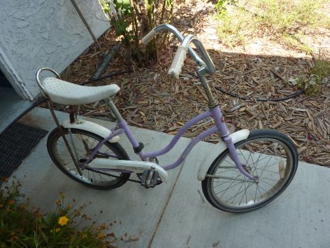 1900 Yugo GVX - A purple bicycle with white fenders and a white banana seat is parked on a concrete surface next to some foliage.