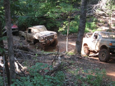 1979 Ford F-150 - Two Ford trucks, one a 1979 F-150, are seen off-roading on a dirt path through the woods.