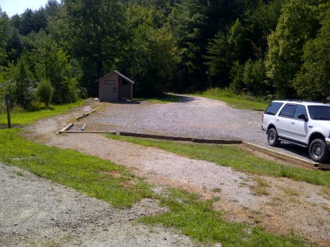 1982 Ford Bronco - A white SUV is parked on a gravel lot surrounded by trees and a small wooden building.