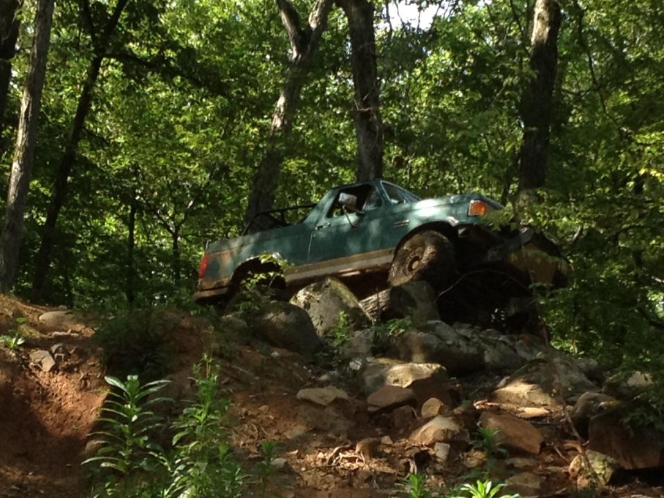 Green pickup truck on rocks, front right tire elevated. Body shows rust. Off-road tires