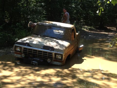 1979 Ford F-150 - A muddy 1979 Ford F-150 is stuck in a water-filled ditch, with people nearby.