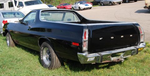 1975 Ford Ranchero - A black 1975 Ford Ranchero is seen from the rear, parked on grass with other vehicles in the background.
