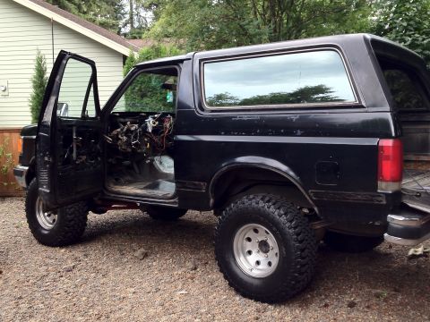 1987 Ford Bronco - A black 1987 Ford Bronco sits with its driver's door open, revealing exposed interior components.