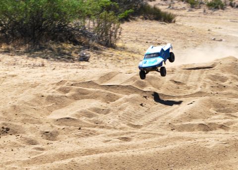 1986 Ford Bronco - A blue remote control off-road truck is captured mid-air as it jumps over a sand dune.