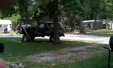 1995 Ford Bronco - A lifted Ford Bronco with camouflage paint sits parked in a grassy area, partially obscured by a tree.