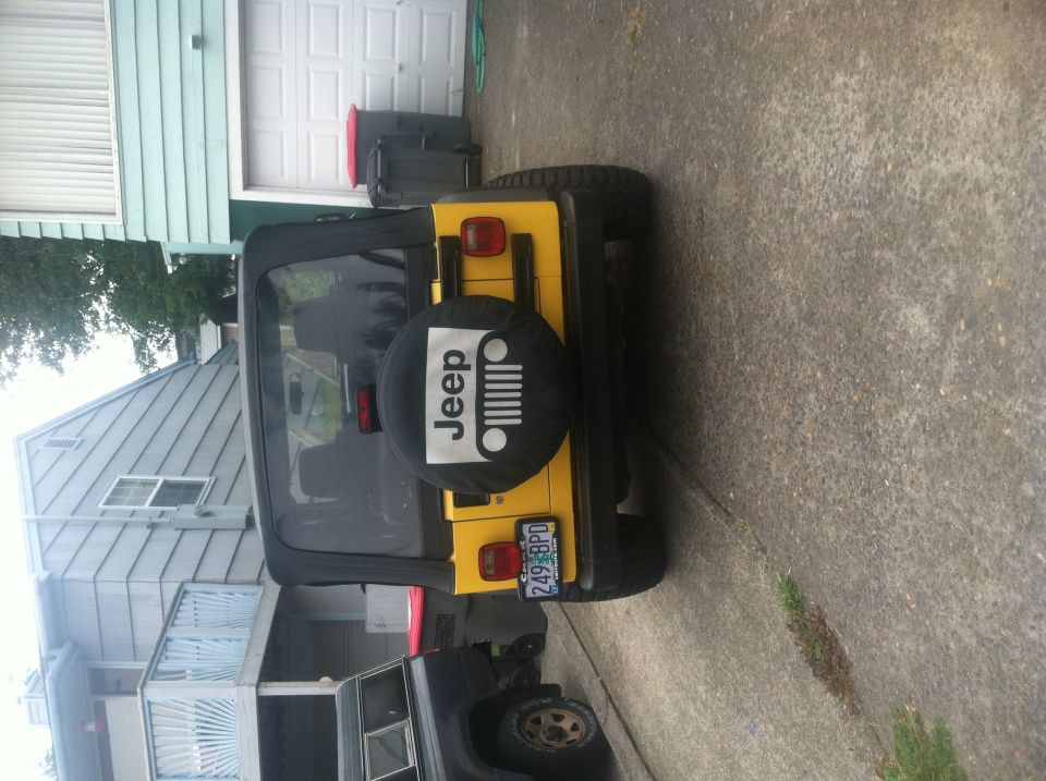 Yellow Jeep TJ parked in driveway, spare tire cover has Jeep logo