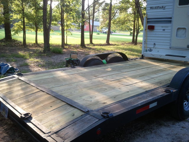 Flatbed trailer with wooden deck, camper hitched to back. Trees and grass in background