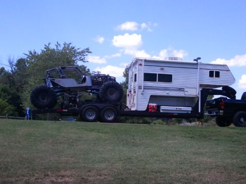 1967 Ford Mustang - Tube-chassis rock crawler buggy on a tandem-axle trailer, towed by a pickup carrying a slide-in camper, parked on grass under blue sky.