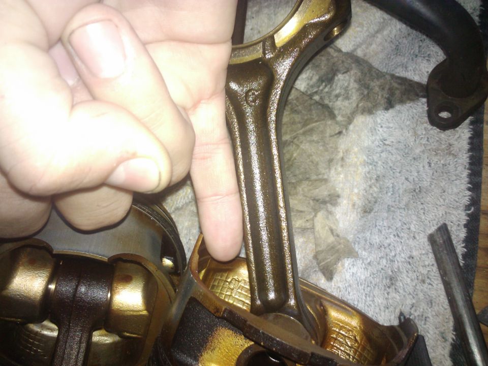 Engine connecting rod held over a piston assembly. A 'G' is visible on the rod