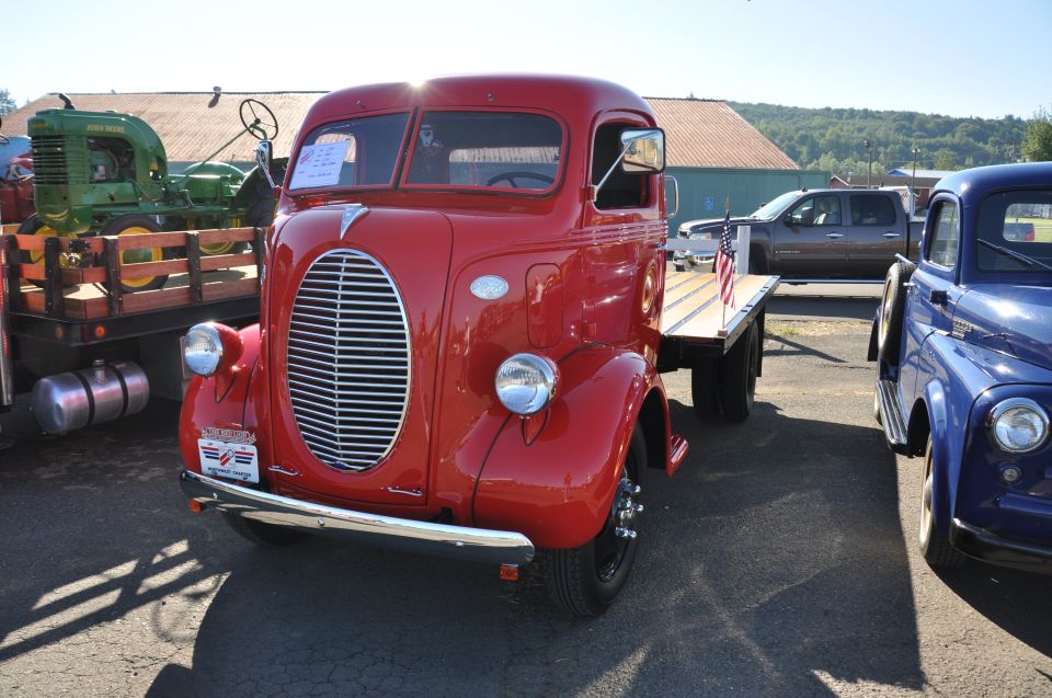 Red flatbed truck with chrome grille and headlights parked next to a blue truck. A green tractor is on a trailer behind the red truck