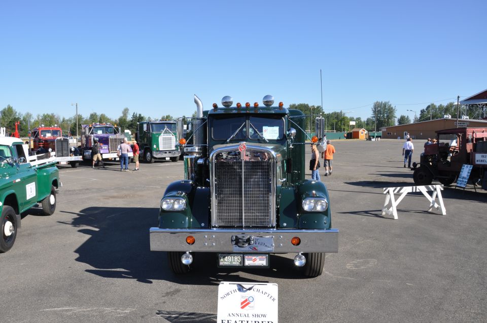 Dark green 1959 Kenworth W900A semi-truck, front view, chrome grille, parked outdoors with other trucks