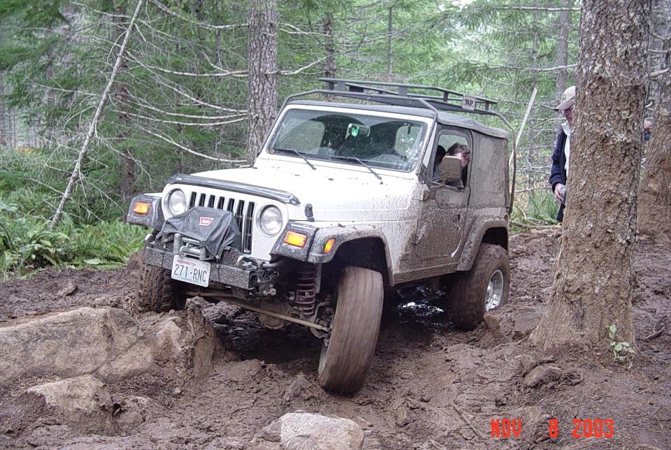 Tan Jeep Wrangler Unlimited muddy on a wet forest trail, roof rack and rear-mounted tire, man standing nearby