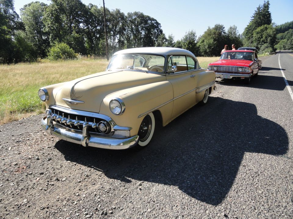 tan 1953 Chevrolet Bel Air parked on gravel next to a red car on the road