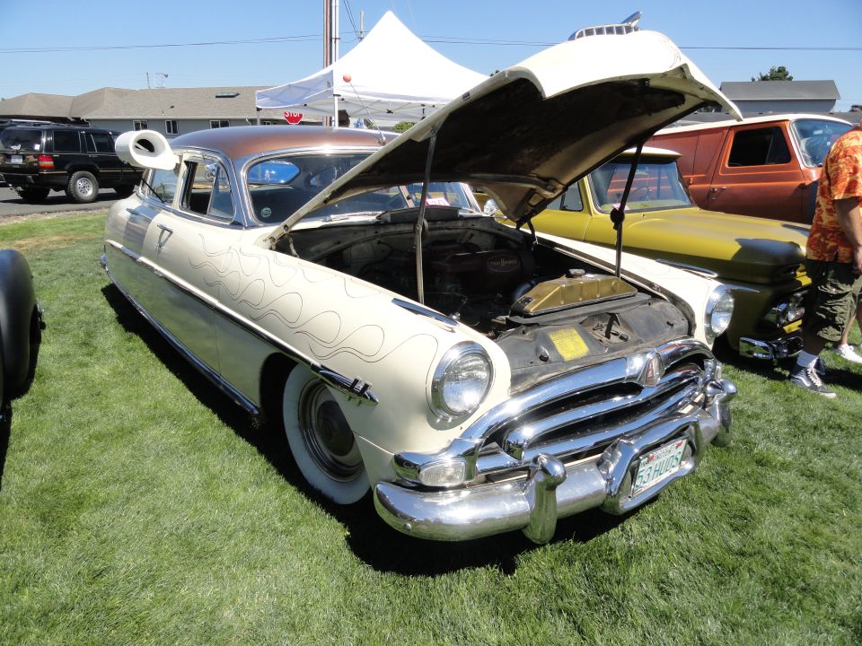 Cream-colored Hudson Hornet with flame decals, hood open, parked on grass next to a yellow truck