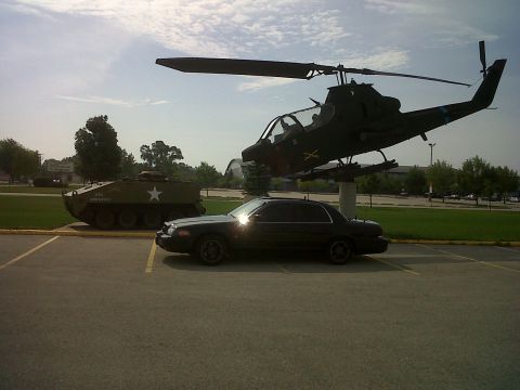 1999 Ford Crown Victoria - A black sedan is parked in a lot with a military helicopter and tank as background elements.