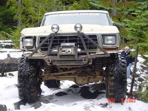 1989 Ford Bronco - A heavily modified Ford Bronco navigates a snowy, rocky trail in a wooded area.