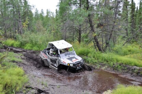 1986 Ford F-150 - A side-by-side off-road vehicle navigates a muddy trail through a dense forest.