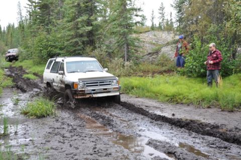 1986 Ford F-150 - A white SUV struggles through a deep mud rut on an off-road trail, with another vehicle in the distance.