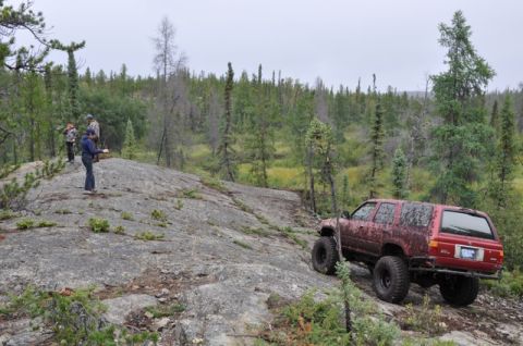 1986 Ford F-150 - A red SUV with oversized tires tackles a challenging rocky incline in a wooded area.