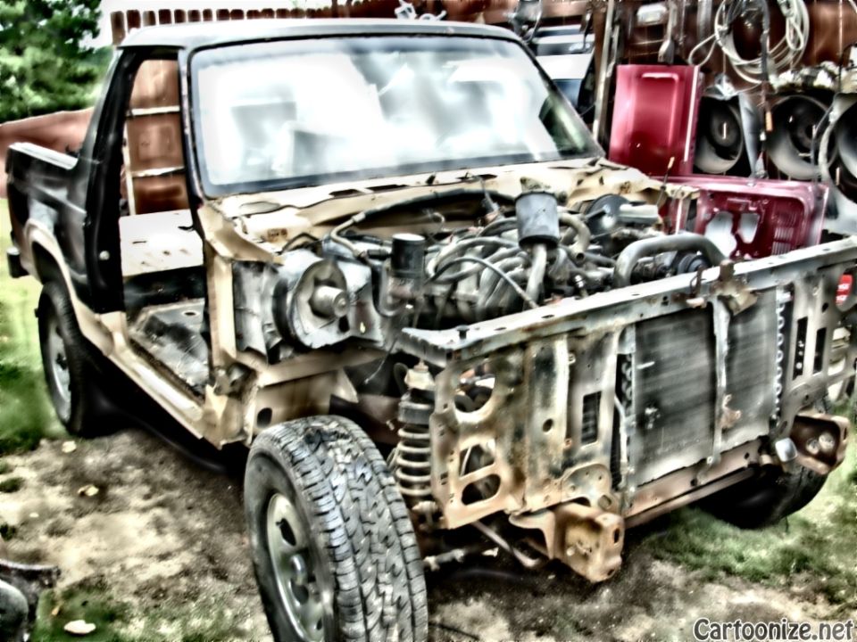 Front-left view of a stripped-down truck with exposed engine and missing fenders in a junkyard.