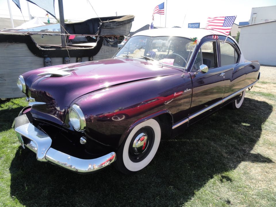 Purple Kaiser sedan with a white roof, parked on grass. American flags are on the roof
