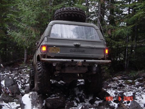 1989 Ford Bronco - An olive green SUV navigates a challenging rocky trail in a wooded area, with a spare tire mounted on its roof.