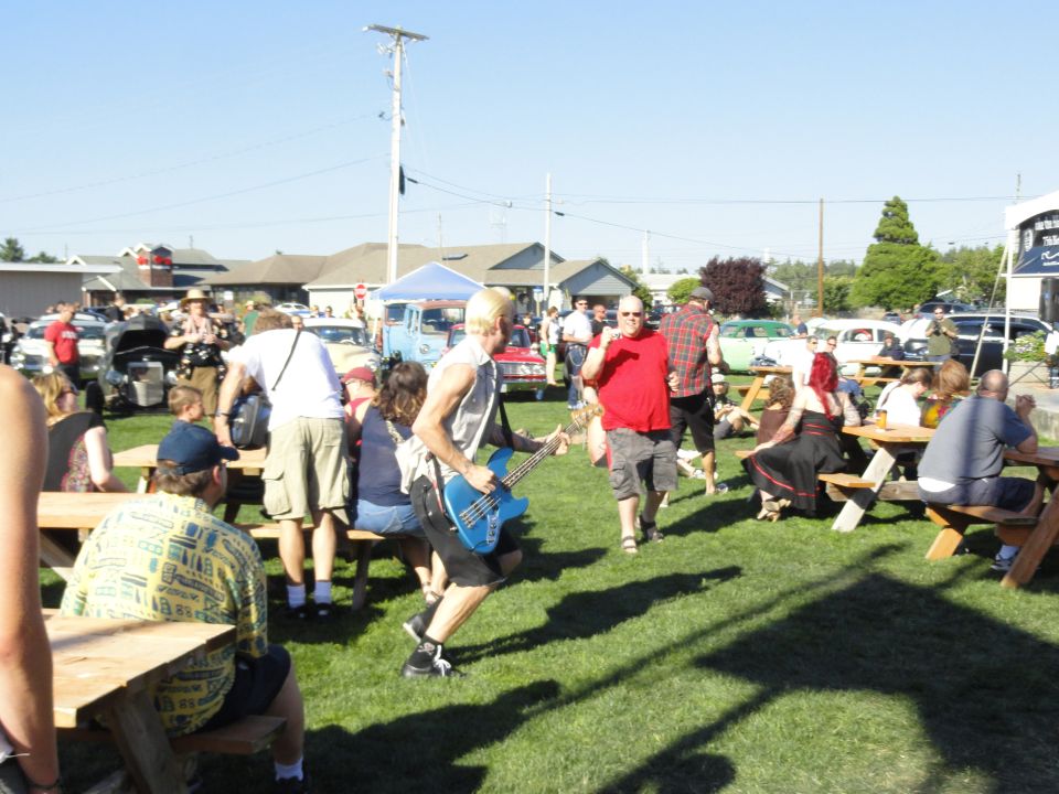 Outdoor event with people gathered around picnic tables and parked cars in the background