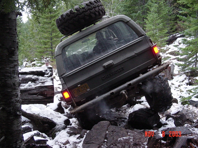 Dark Chevrolet SUV with roof spare climbing snowy rocks on a wooded trail, seen from behind