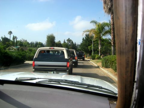 1996 Ford Bronco - A line of vehicles drives down a suburban street on a sunny day, with palm trees and houses visible.
